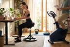 Kvinde arbejder ved et skrivebord på en ergonomisk stol, mens en mand bruger en motionscykel i et lyst hjemmekontor.